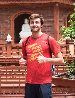 Joey Reilman stands and smiles at the camera holding two thumbs up in front of a religious East Asian shrine.