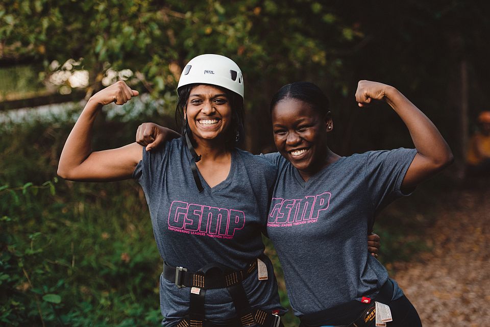 Sangee and Caroline flex their muscles and smile at the camera. Sangee is wearing a helmet used for zip lining.