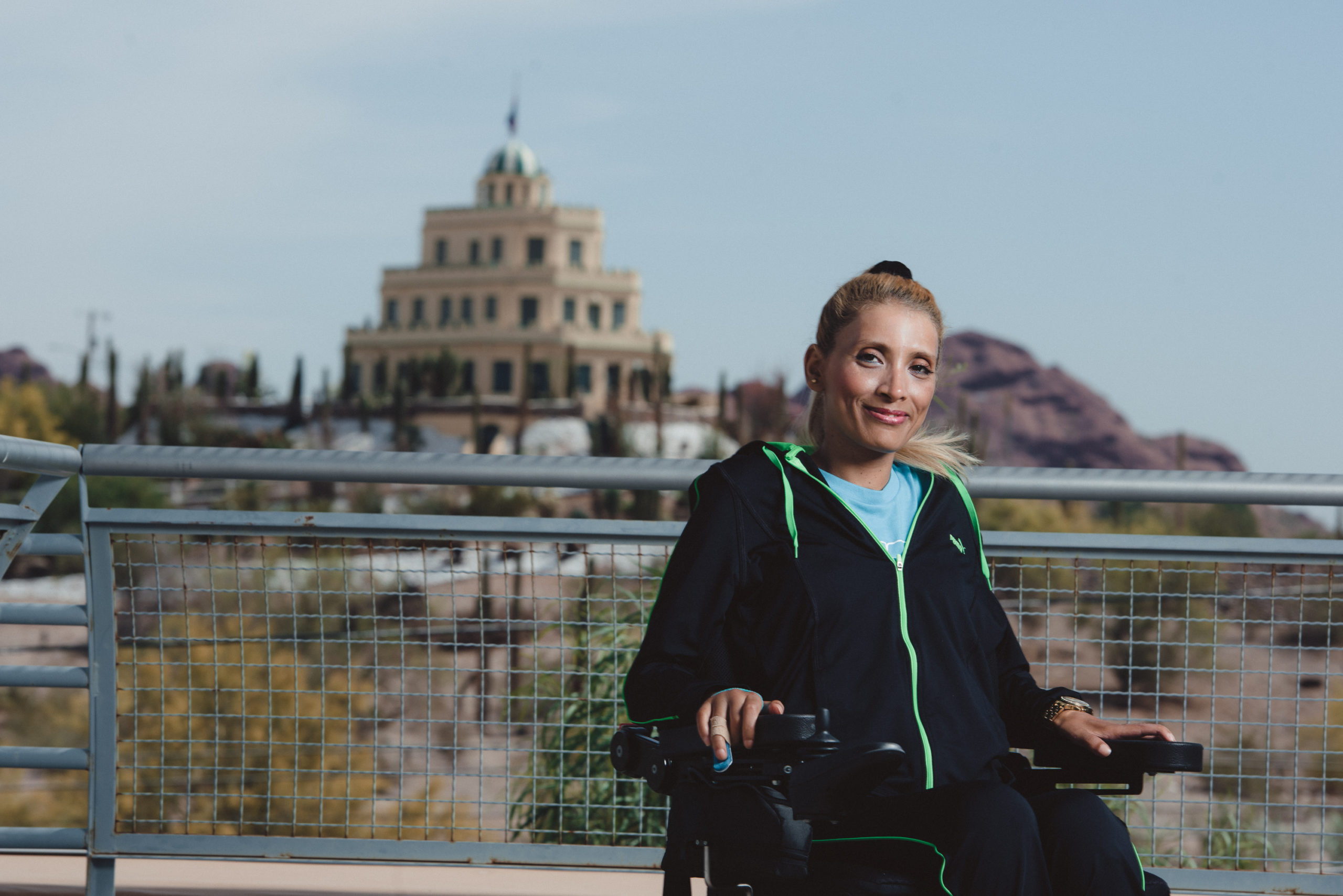 Rola Al-Amer Allaweh sits in her wheelchair smiling at the camera. In the background is a four story structure in the desert of Arizona.