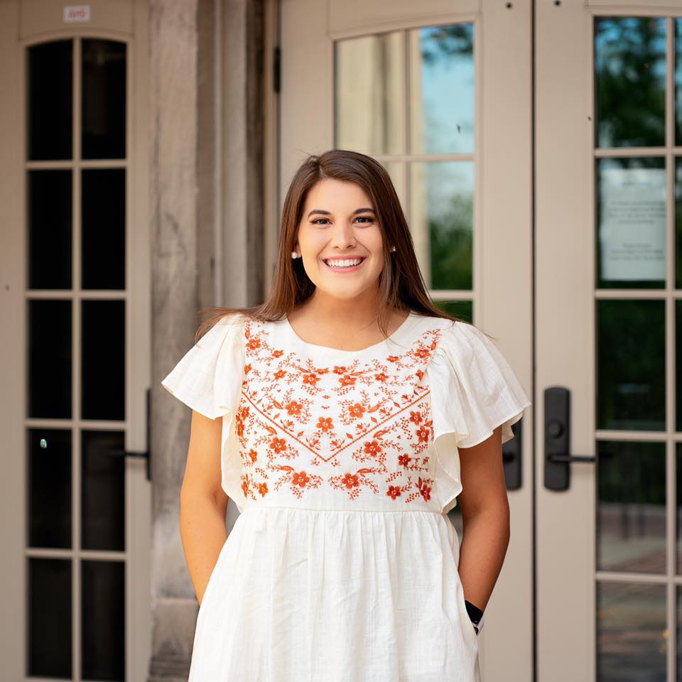 Camryn Cupp stands smiling at the camera with her hands in the pockets of a white dress.