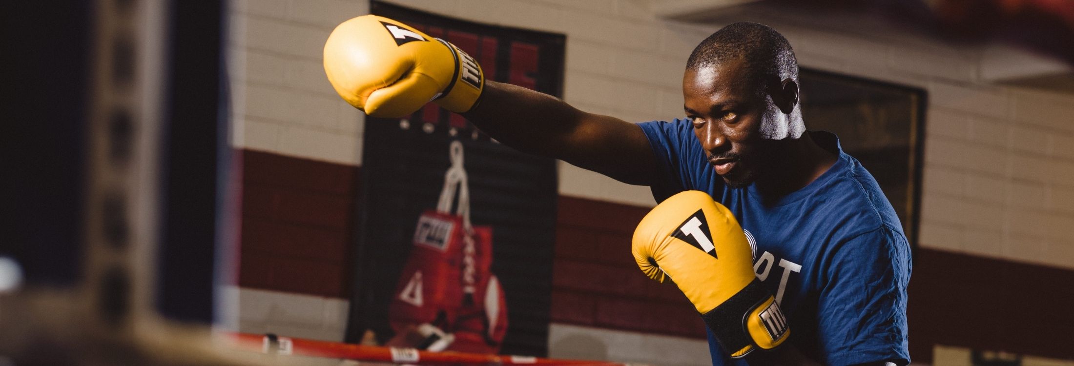 Hassan Badjdi, a 2019 Sport 4 Community participant from Senegal, stands looking to the left wearing a navy blue shirt, posing in a straight punch stance wearing bright yellow boxing gloves.